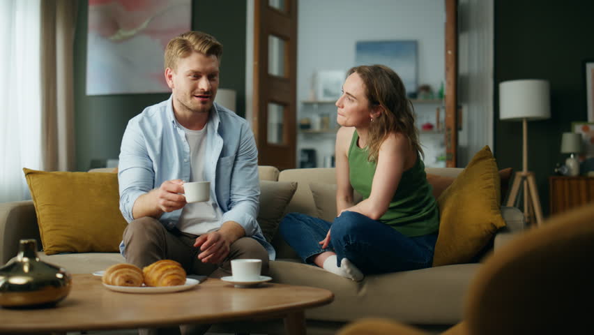 Chilling spouses drinking tea at sofa interior. Love couple enjoying coffee cups talking together at apartment. Smiling husband wife sipping hot beverage relaxing couch. People home lifestyle concept Royalty-Free Stock Footage #1102818239
