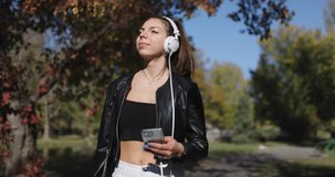 Beautiful girl with headphones looking at phone. Fashionable young lady walking in the park. Slow motion handheld video recorded at 120fps.