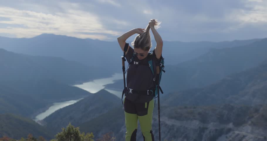 Active girl with hiking poles and backpack enjoying a view of river lake from high in the mountains. Standing on a cloudy and windy weather.