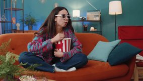 Excited young woman in 3D glasses sits on sofa eating popcorn snacks and watching interesting TV serial sport game film online social media movie content at home apartment. Girl enjoying entertainment