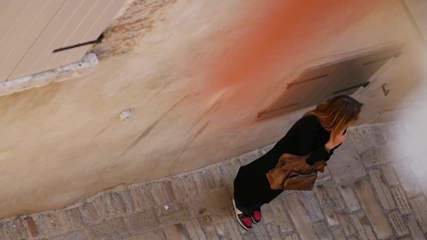 Young beautiful woman stands on street of old city and calls on phone. Tourist is trying to find address while standing on street. View from above. Sunny day. Rovinj, Istria, Croatia - April 10, 2023