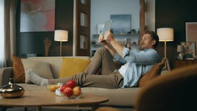 Relaxed man calling tab laying sofa. Positive hipster holding tablet computer reacting at interlocutor story at cozy apartment. Modern blogger recording content at home. Guy making video conference - Powered by Shutterstock - Get 15% off with code: PIKWIZARD15
