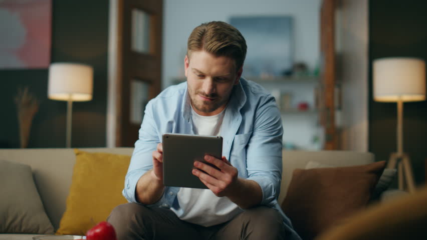 Positive man using tab computer at home. Optimistic guy working pad sitting sofa in living room. Relaxed businessman holding digital tablet browsing online. Smiling freelancer examining file remotely  Royalty-Free Stock Footage #1102856307