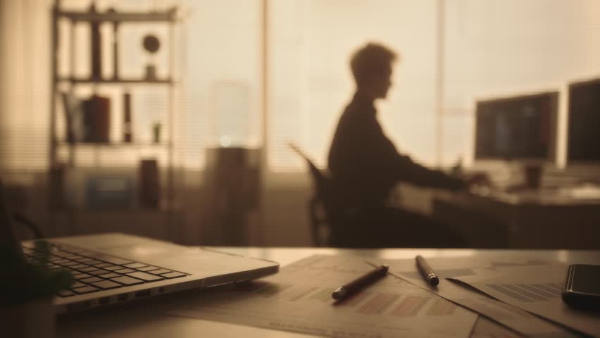 Blurred silhouette of a young man typing on a computer keyboard and drinking coffee in a dark office in the evening. Manager works sitting at a table near a window with backlight. Royalty-Free Stock Footage #1102863077