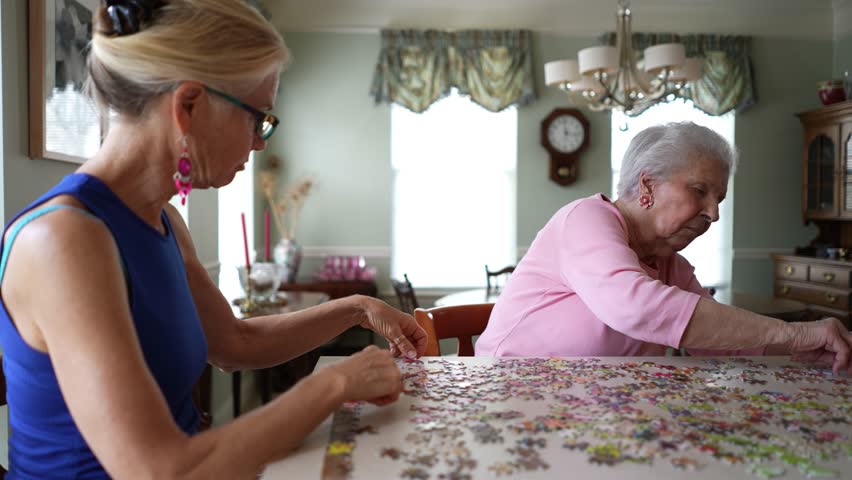 Camera moving around table as two happy elderly senior woman sitting at table working on puzzle with mature middle aged daughter.