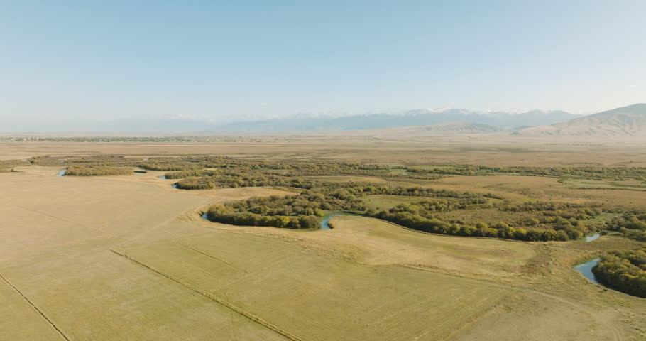 Curving flat Bayankol River in Kazakhstan with small oasis around among arid dry fields. Mountainous range background with snow mount peaks. Drone view at autumn sunny sunset. Royalty-Free Stock Footage #1102868969