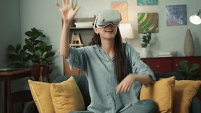 Young Beautiful Asian woman putting on Virtual Reality headset for first time exploring VR world playing game with curious expression pointing fingers on screen sitting on sofa in cozy living room