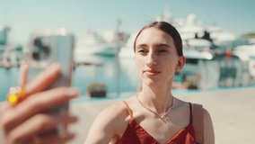 Сlose-up young woman takes a selfie, video call sitting at the seaport uses a mobile phone. Smiling girl Use Social Media, Streaming Service in the background yachts and ships