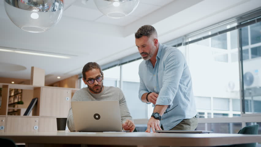 Two software developers discussing a project in an office, using a laptop to review their progress. Innovative business men collaborating on a tech solution using their expertise in the field.