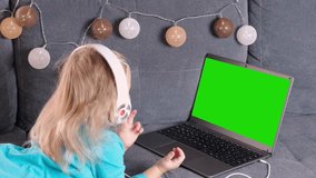 child 2-3 years old, girl in headphones lies on sofa in front of laptop, uses Internet, watches educational cartoons, plays computer games, green blank screen for video, concept of remote learning