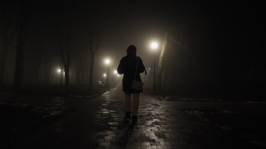 A woman looks around cautiously walking through a city park in the late evening Royalty-Free Stock Footage #1102888115