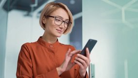 Close up of a happy young blonde woman in glasses using a smartphone in a modern office. Smiling stylish female employee typing a message, chatting online with a friend or browsing social media - Powered by Shutterstock - Get 15% off with code: PIKWIZARD15