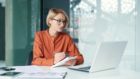 A young woman is taking online video conferencing training using a laptop while sitting in a modern office. Concentrated female in glasses remotely communicates with coach and writes notes in notebook - Powered by Shutterstock - Get 15% off with code: PIKWIZARD15