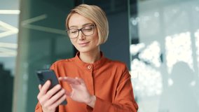 Young female employee wearing glasses is using smartphone while sitting at workplace in modern office. Smiling blonde woman typing message, chatting online, browsing social media or checking e-mail - Powered by Shutterstock - Get 15% off with code: PIKWIZARD15