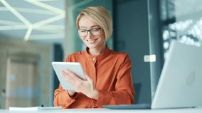 Happy young blonde woman using tablet while sitting at workplace at desk in modern office. Smiling female employee wearing glasses browses social networks, reads email messages, chats online - Powered by Shutterstock - Get 15% off with code: PIKWIZARD15
