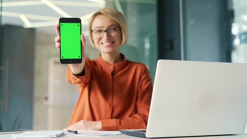 Young smiling woman showing smartphone with green screen while sitting at workplace in modern office. Color key vertical mockup. Template for advertising. Happy female employee looking at camera - Powered by Shutterstock - Get 15% off with code: PIKWIZARD15