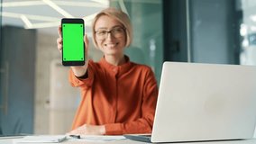 Young smiling woman showing smartphone with green screen while sitting at workplace in modern office. Color key vertical mockup. Template for advertising. Happy female employee looking at camera - Powered by Shutterstock - Get 15% off with code: PIKWIZARD15