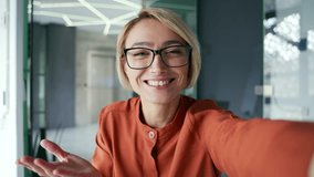 Webcam view. Yung smiling woman is having a video call using a smartphone while sitting at a workplace in a modern office. Happy female employee looking at camera, waving, laughing, talking to friend - Powered by Shutterstock - Get 15% off with code: PIKWIZARD15