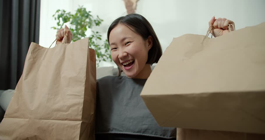 Excited asian lady shopping online, lifting gift bags above her head at home