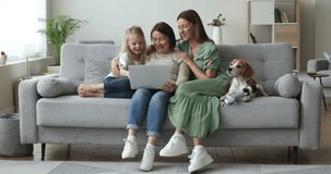 Grandmother, her daughter, little granddaughter and cute dog relax on sofa in living room, spend weekend together at home on internet, use laptop to watch videos, do e-shopping via ecommerce website