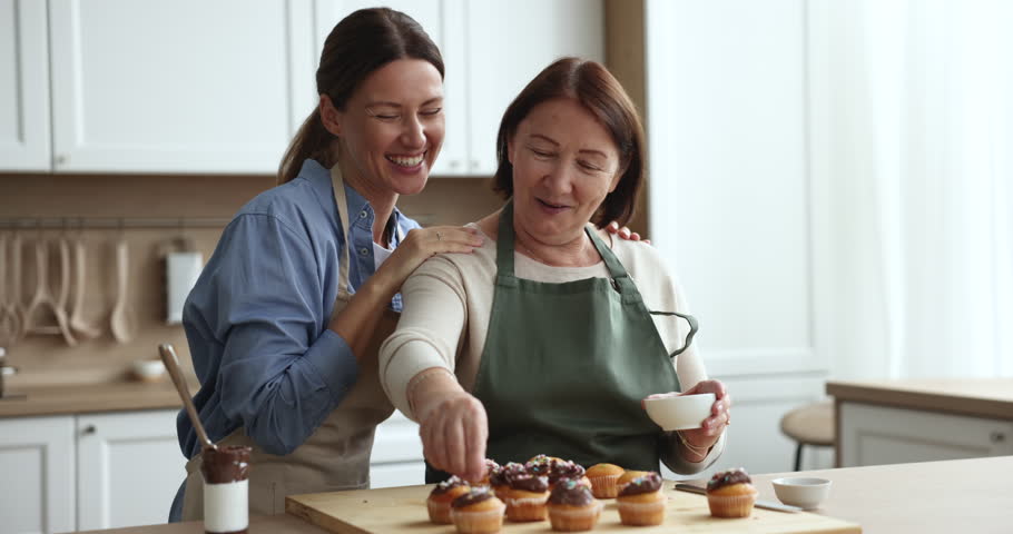 Young adult 35s daughter and aged mom wear aprons prepare dessert, talking, hugging, sprinkles fresh baked cupcakes with sugar topping, cooking in cozy domestic kitchen enjoy pastime and time together | Shutterstock HD Video #1102892563