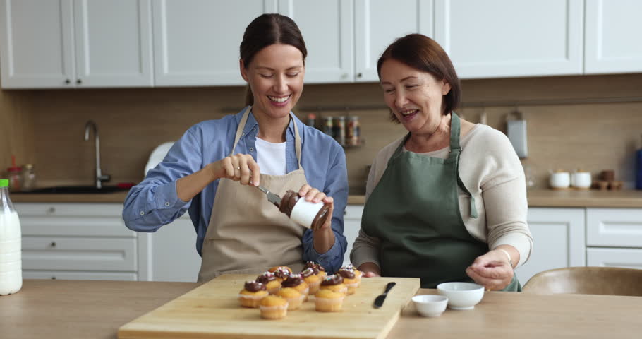 Grown up daughter and older mother wear aprons prepare dessert, talking, hugging, decorate cupcakes with chocolate flavored paste cook together in cozy domestic kitchen enjoy pastime and communication Royalty-Free Stock Footage #1102892565