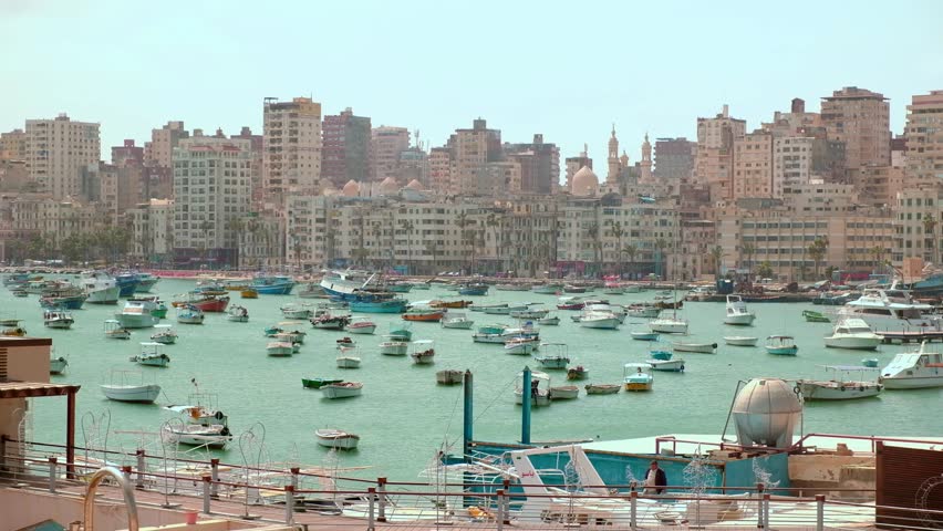 Fishing boats and dinghies moored in Alexandria harbor. Yachts and fishermen boats rocking on sea waves in a harbor opposite Corniche, residential buildings and mosque at background. Alexandria, Egypt
