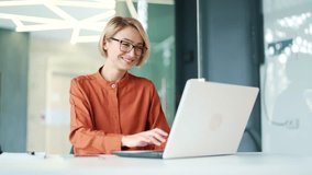 Happy young employee wearing glasses typing on laptop while sitting at workplace in modern office. A smiling blonde female chats online, communicates with a colleague, a friend, replies to a client - Powered by Shutterstock - Get 15% off with code: PIKWIZARD15