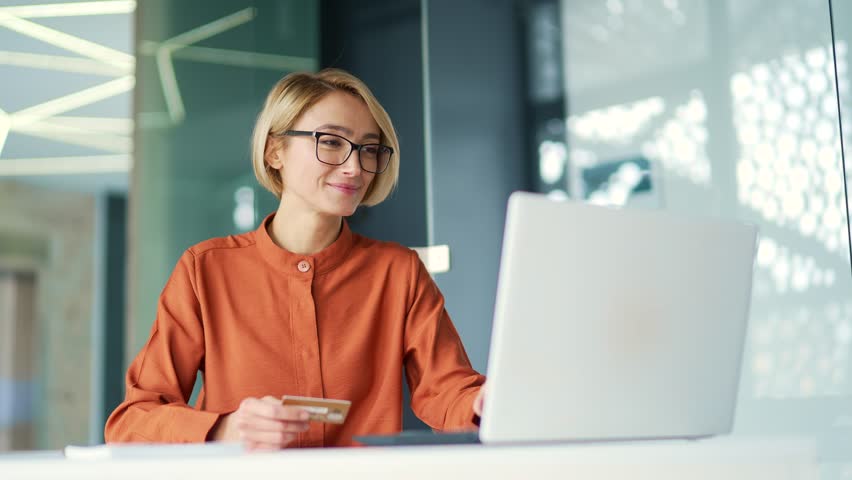 Happy young blonde woman doing online shopping typing credit card number on laptop while sitting at desk in modern office. A smiling and satisfied female employee in glasses happily makes a purchase Royalty-Free Stock Footage #1102895917