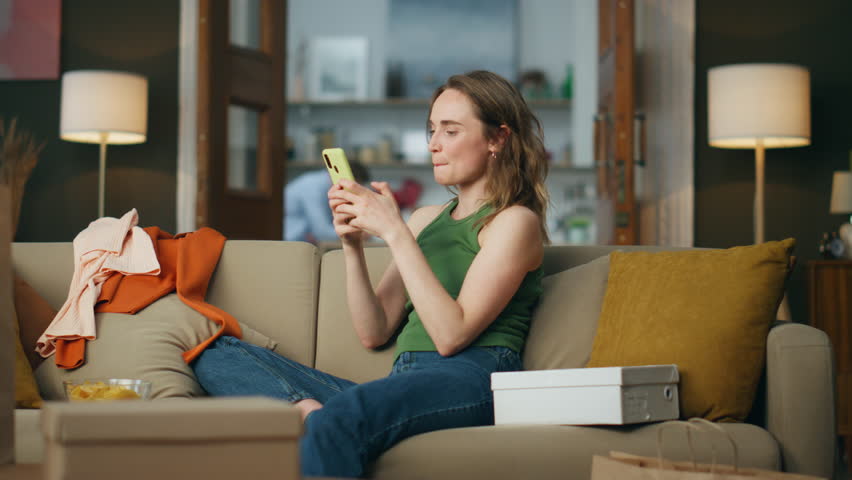 Emotional girl rejoicing mobile phone at home. Shopping addicted person sitting sofa ordering new items. Happy smiling woman dancing hands with smartphone relaxing around boxes and clothes alone Royalty-Free Stock Footage #1102897731