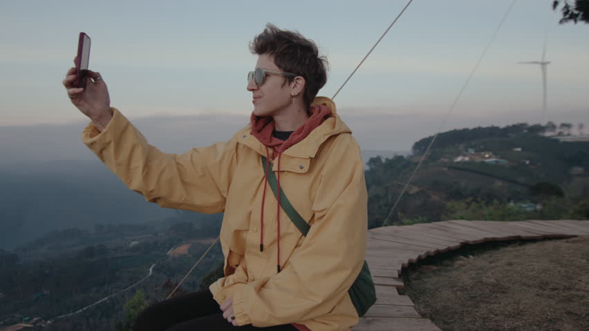 Young female tourist sitting at the viewpoint on mountain peak, holding smartphone and taking photos of scenic nature around, smiling and enjoying weekend getaway