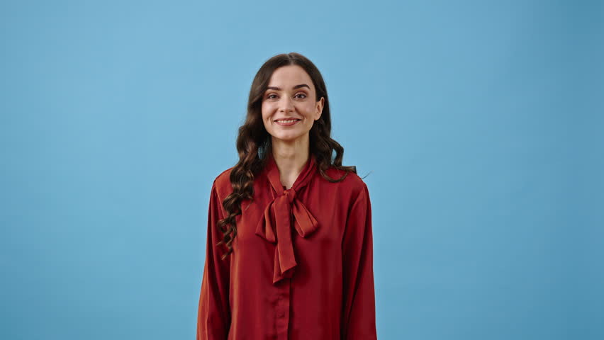 Smiling young woman shows Okay sign. Pretty female in crimson silk clothes demonstrates agreement and support. Positive emotions on blue background Royalty-Free Stock Footage #1102906223