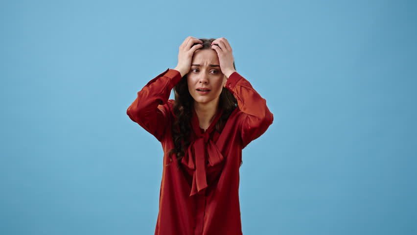 Frightened young woman covers mouth with palm in shock. Upset female holds head in hands and looks around with nervous expression on blue background
