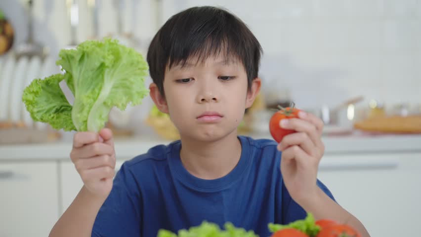 Asian little boy frowning unhappy and don't like eating vegetables holding greens vegetables and tomato on the tabel at kitchen room. Nutrition  healthy eating habits for kids concept | Shutterstock HD Video #1102912997