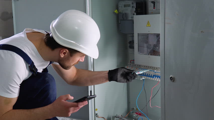 Handsome bearded electrician repairing electrical box. Electrician using screwdriver and a new application on a smartphone Royalty-Free Stock Footage #1102917781