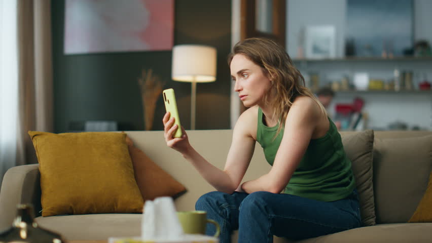 Unhappy woman looking telephone screen at living room. Sad girl getting bad news holding smartphone on sofa. Worried lady checking phone feeling depressed in modern house. Dismal person sighs heavily Royalty-Free Stock Footage #1102919281