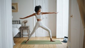 Flexible woman stretching body in cozy place. Curly hair athlete practice asana on yoga carpet. Yogi model watching video tutorial using tablet. African girl training alone. Harmony lifestyle concept 