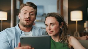 Shocked couple staring pad digital device at home closeup. Amazed spouses examining tablet computer screen at house zoom out. Bearded wondering man holding gadget laughing at cozy living room interior - Powered by Shutterstock - Get 15% off with code: PIKWIZARD15