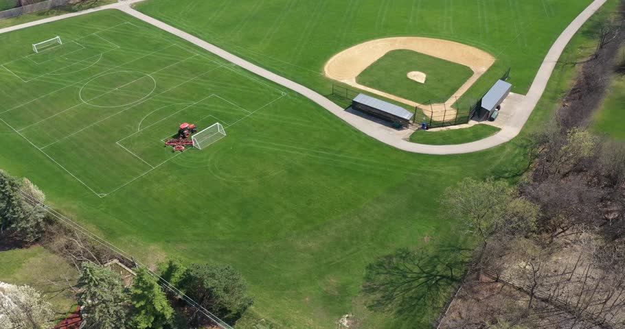 Aerial view of a baseball and soccer field in a large suburban park with a sports field mower tractor grooming the grass in early spring. Royalty-Free Stock Footage #1102922227