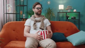 Excited young man in 3D glasses sits on couch eating popcorn snacks and watching interesting TV serial sport game, film online social media movie content at home apartment. Guy enjoying entertainment