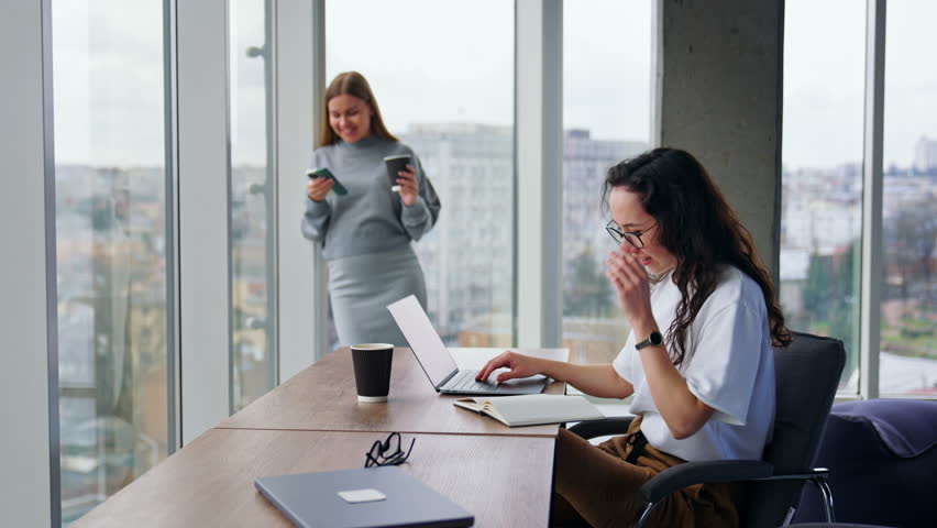 Dark-haired woman finishes typing on laptop, puts off glasses and shows her paper notebook to colleague. Teammates look at notebook, discussing issues in it.