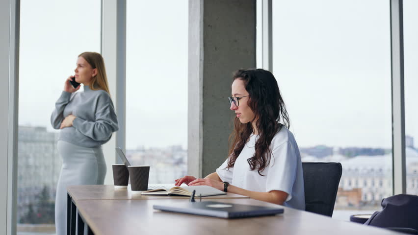 Business people working in the light office. Brunette lady works on laptop at desk and blonde pregnant woman speaking on the phone.