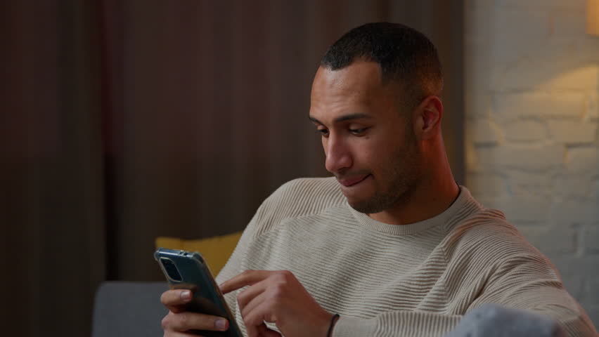 Multiracial married couple use mobile phone at home smiling muslim woman wife girl in hijab and african american man husband guy surfing smartphone internet shopping online make order web store app