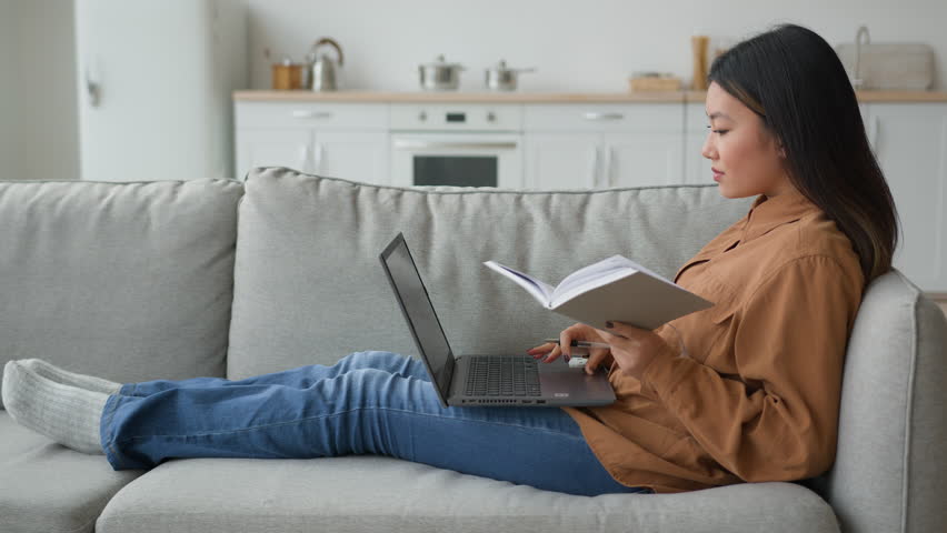Side view Asian woman girl on sofa with laptop student writing notes in copybook preparing for exam doing homework working from home watching educational lesson studying online search data on internet Royalty-Free Stock Footage #1102935009