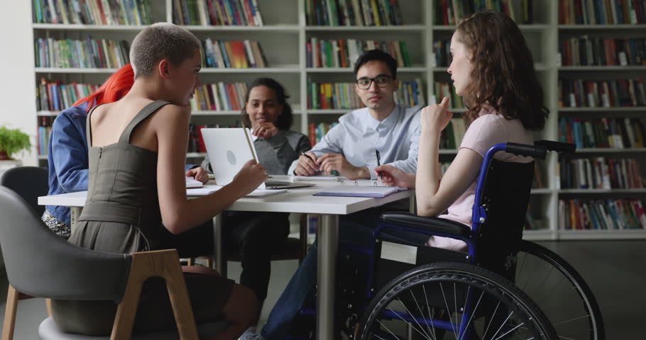 Teen schoolgirl sits in wheelchair take part in group meeting with multi ethnic groupmates discuss joint assignment gathered in library. Accessibility of education of person with physical disability Royalty-Free Stock Footage #1102943903