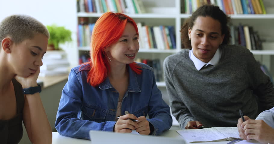 Pretty girl with dyed bright red hair talk to schoolmates about joint task, share ideas, creative vision sit at desk in library or classroom. Teamwork, education, companionship of multiethnic students