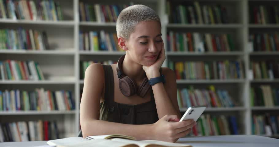Pretty young girl distracted from studying in library use modern smartphone enjoy chat, remote communication with friend, check timetable or grades through mobile app smile feel satisfied. Technology Royalty-Free Stock Footage #1102944703