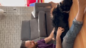 Young man sitting on the living room floor playing with his young female dog (Australian shepherd). Vertical video 4K.