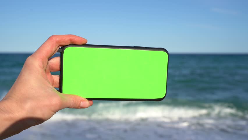 Hand holding horizontal cell mobile phone with green chromakey screen in front of sea waves. Template mock up video footage. Nature background