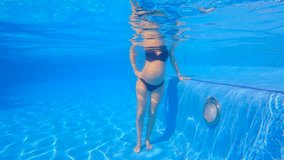 In this slow-motion underwater video, a pregnant woman is seen performing water exercises in a swimming pool. With her baby bump, she moves with ease in the water, demonstrating the benefits of